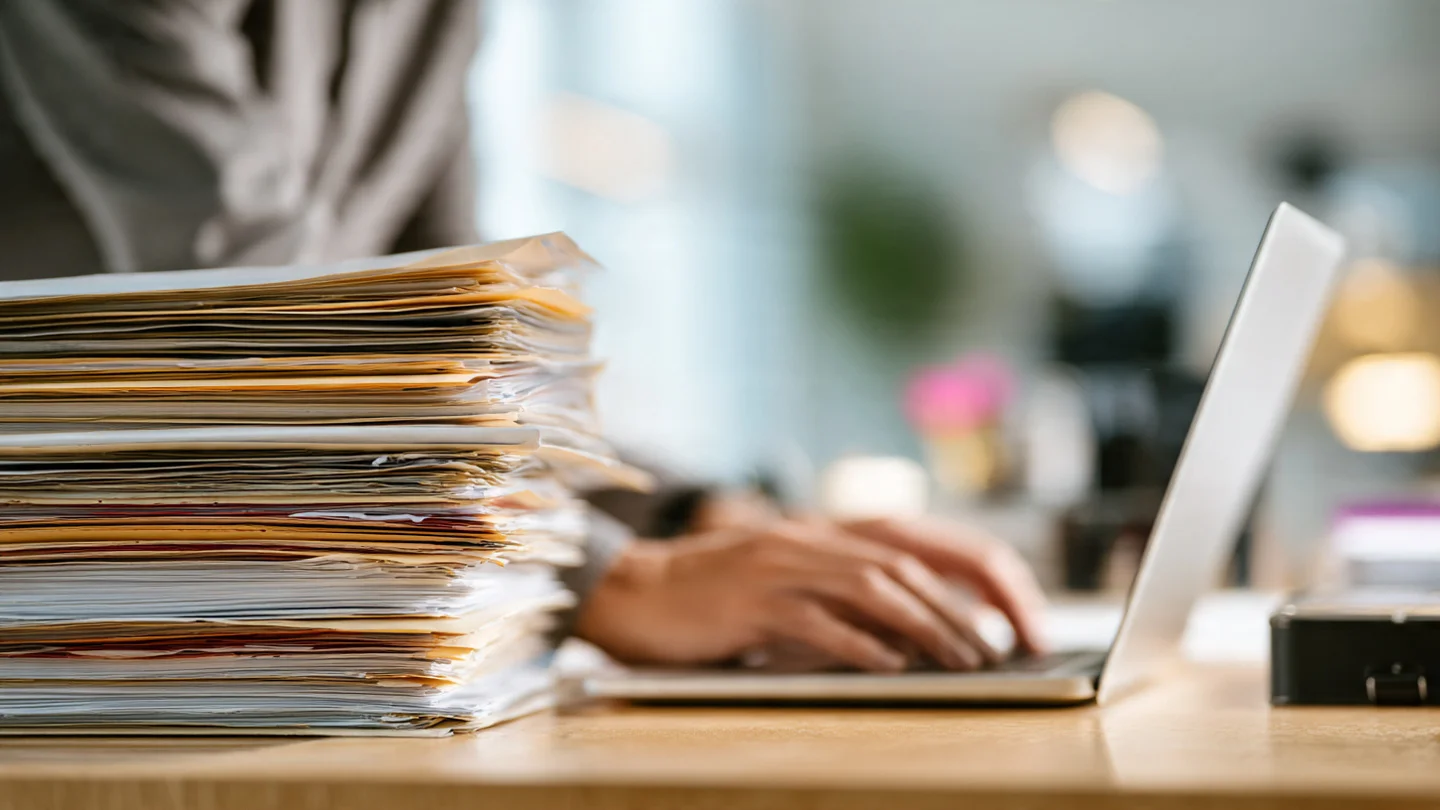 A person typing on a laptop next to a large, organized stack of paper documents and folders.